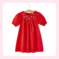 A bright red short-sleeve baby dress with puff sleeves and white embroidered flamingos on the yoke, hanging on a wooden hanger against a white background with a soft pink border.