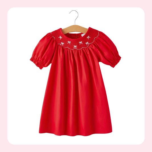 A bright red short-sleeve baby dress with puff sleeves and white embroidered flamingos on the yoke, hanging on a wooden hanger against a white background with a soft pink border.