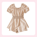 A light beige linen romper with short puff sleeves, a smocked waist, and a square neckline, displayed on a white background with a soft pink border.