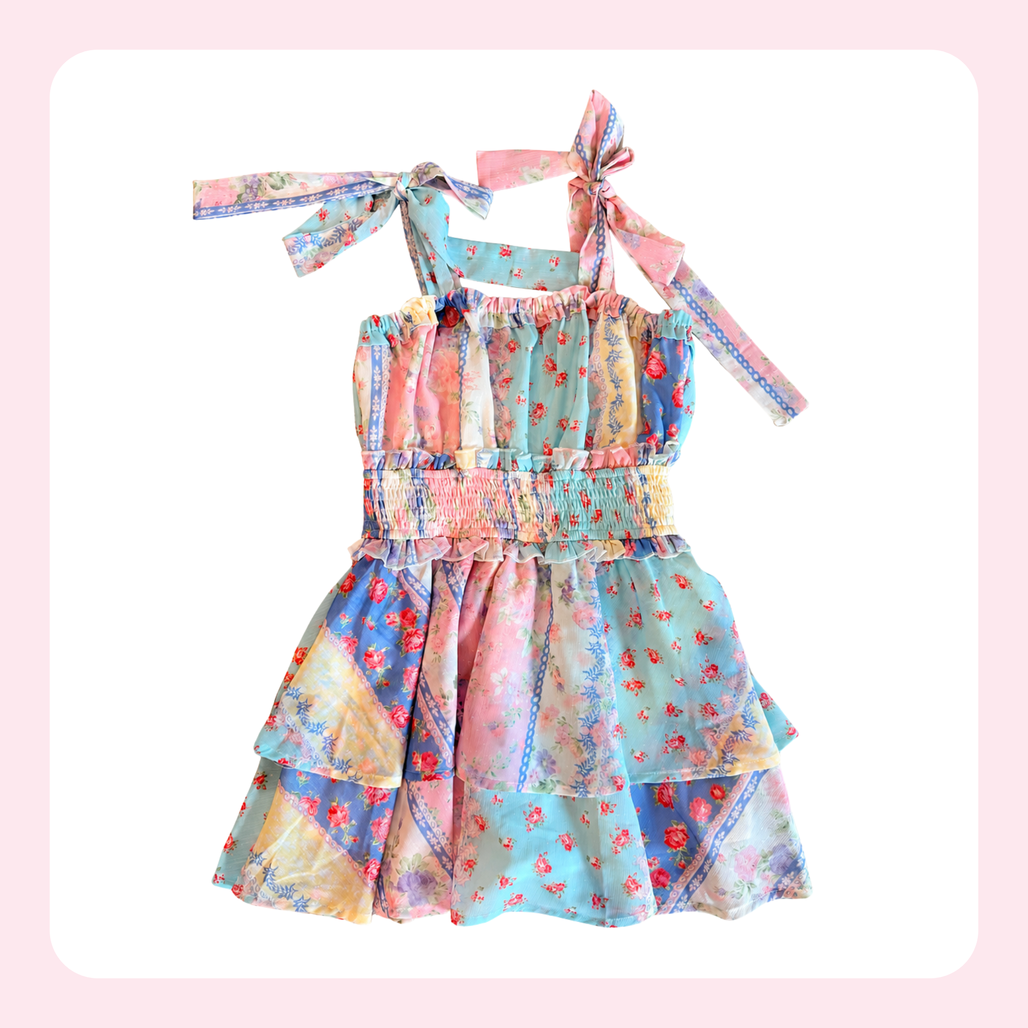 A sleeveless tiered dress with a colorful patchwork floral print in pastel shades of blue, pink, and yellow, featuring tie shoulder straps and a smocked waist, shown on a white background with a soft pink border.