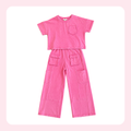 A two-piece children's outfit in a bright pink washed cotton fabric, featuring a short-sleeve cropped T-shirt with a chest pocket and matching wide-leg cargo pants with front and side pockets, displayed flat against a white background with a light pink border.