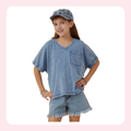 A young girl modeling a light blue acid-wash denim two-piece set, featuring a short-sleeve V-neck t-shirt with a chest pocket and matching frayed-hem denim shorts, styled with a matching denim cap against a white background with a pink border.