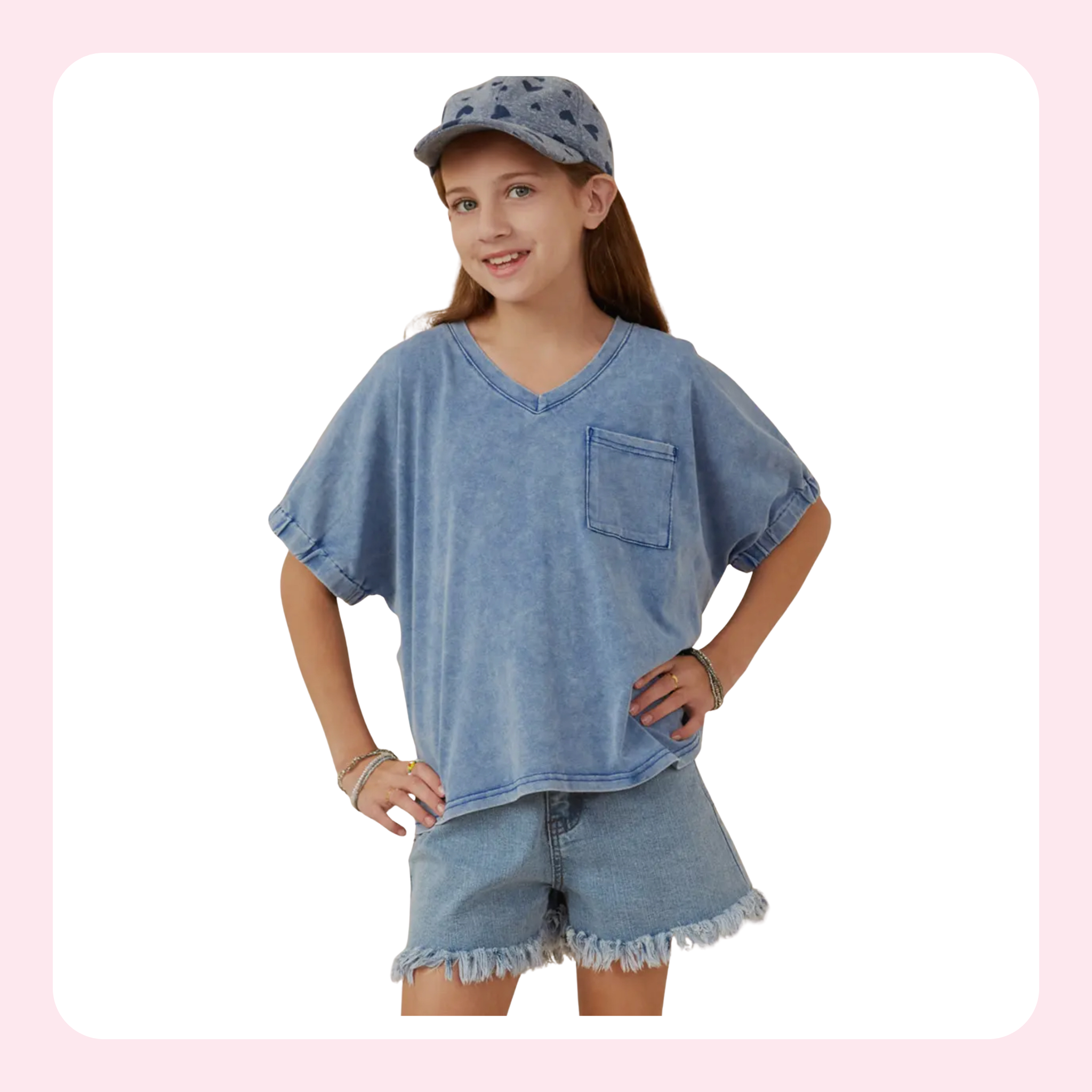 A young girl modeling a light blue acid-wash denim two-piece set, featuring a short-sleeve V-neck t-shirt with a chest pocket and matching frayed-hem denim shorts, styled with a matching denim cap against a white background with a pink border.